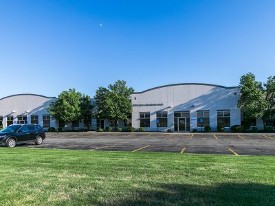 Exterior of a light-gray industrial warehouse with curved roofs, trees in front, and a parking lot with a car under a blue sky.