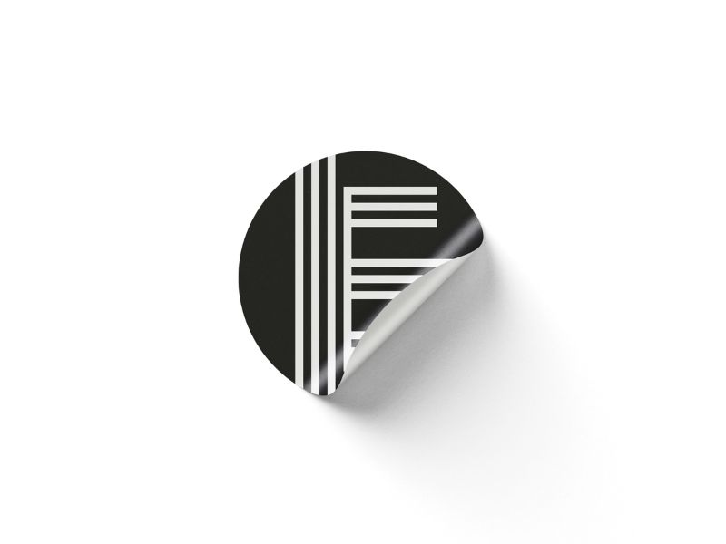 Round black sticker with white vertical and horizontal lines; lower-right corner curls upward, revealing a white surface outside the sticker.