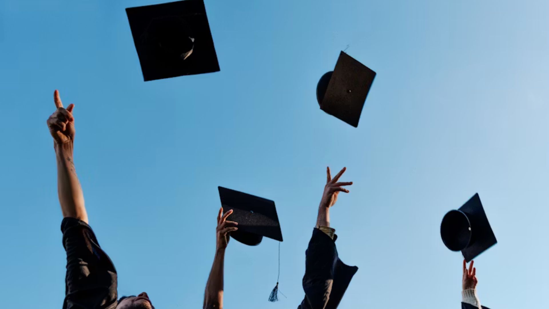 Graduates tossing black mortarboard caps into the air against a clear blue sky, celebrating graduation.