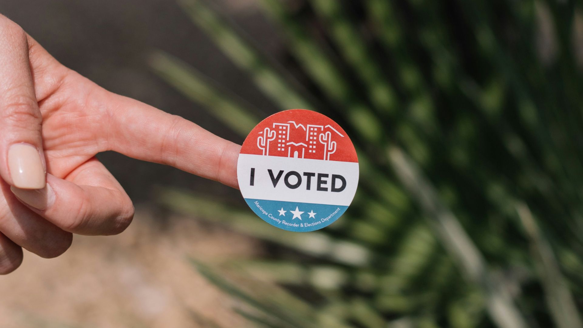 A hand holding a red-white-blue "I Voted" sticker with a desert town graphic at the top and stars at the bottom.
