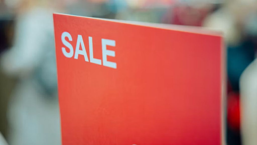 Red sale sign with white 'SALE' text in focus against a blurred store backdrop.